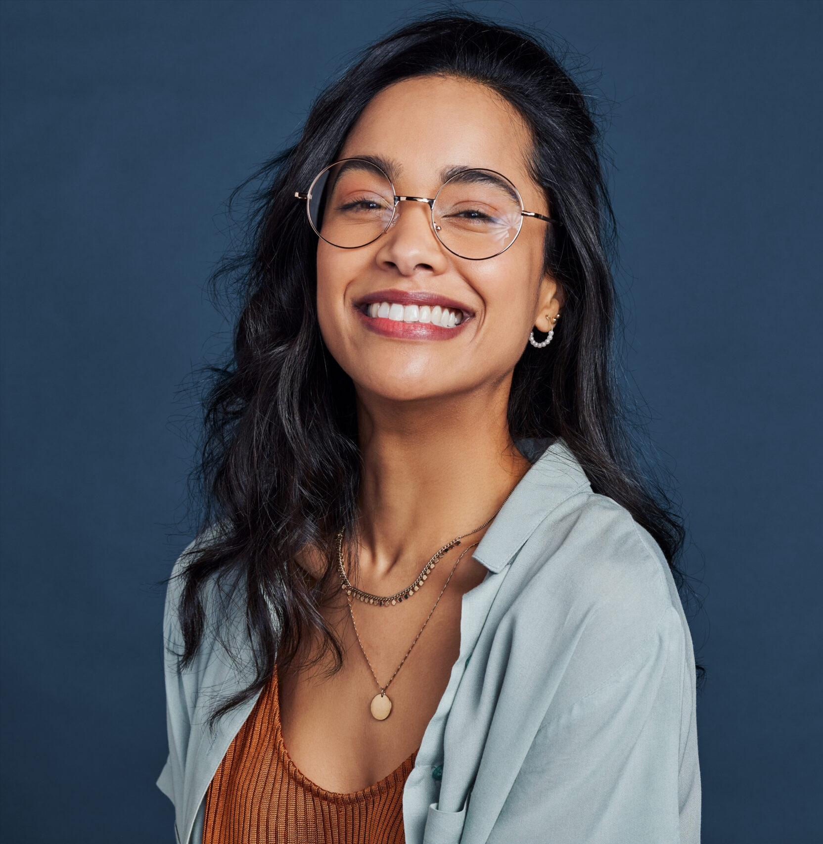 Casual young multiethnic woman with eyeglasses smiling at camera on blue background. Close up face of happy mixed race girl laughing with eyeglasses isolated with copy space. Beautiful university student having fun isolated against blue wall while wearing specs.